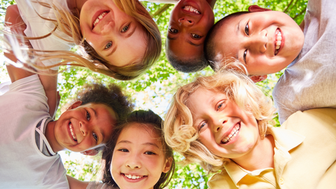 Group of diverse, smiling children huddled in a circle outdoors, looking down at the camera