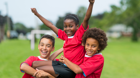 Three children in matching sports uniforms celebrate together on a field, showing teamwork and joy