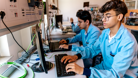 High school students in lab coats work on desktop computers during a hands-on technical training class