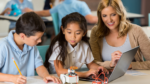 Teacher guiding two students as they work with a small robot and laptop during a hands-on learning activity